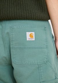 Jasnozielone spodnie cargo z wyraźną tylną kieszenią, z białą metką z pomarańczowym logo i czerwonym tekstem, wykonane z trwałego materiału.