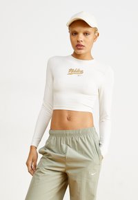 Baltas ilgas rankovėmis trumpas viršus su auksiniu "Nike" logotipu, derinamas su šviesiai žaliais, laisvai krentančiais kelnėmis ir atitinkančia kepure. Visame paviršiuje yra glotni tekstūra.