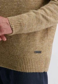 Mann in beigem Wollpullover mit gerippten Bündchen und Saum, einem kleinen schwarzen rechteckigen Markenetikett nahe dem Saum, kombiniert mit dunkler Hose.