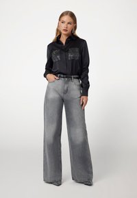 Zwarte satijnen shirt met geborduurde patches, gecombineerd met lichtgrijze high-waisted wijdse spijkerbroeken en gestreepte zwarte hakken.