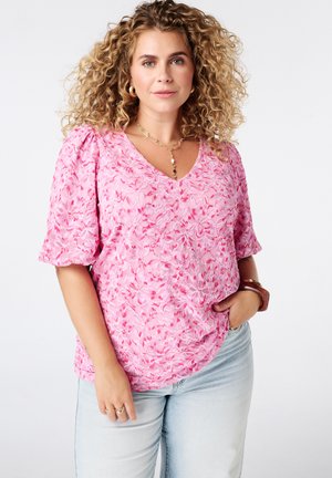 Femme aux cheveux bouclés porte un haut en dentelle florale rose, un jean clair, des bijoux en or et un bracelet marron, debout avec une main dans la poche.