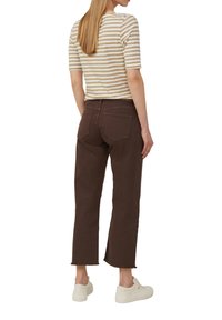 Brązowe spodnie o kroju 7/8 z surowym wykończeniem, zestawione z pasiastą beżowo-białą koszulą, widok z tyłu. Materiał wydaje się być bogaty w bawełnę.