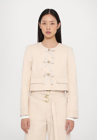 Cropped beige jacket with four front buttons, textured fabric, and ribbon accents. Paired with high-waisted beige pants featuring gold hardware.