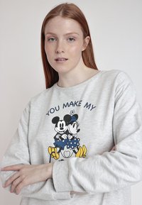 Sudadera de color gris claro con mangas largas que presenta un gráfico frontal de Mickey y Minnie Mouse, el texto "TÚ HACES MI" y acentos coloridos.