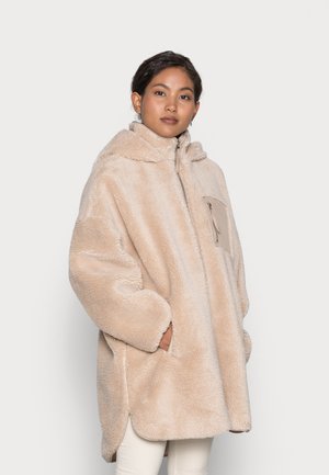 Beige nepbont oversized jas met capuchon, ritsluiting en voorzak. Zachte textuur en ronde randen. Lange mouwen en zijsplitten.