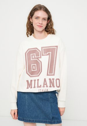 Ung kvinde iført hvid sweatshirt med stort "67 MILANO ITALIA" tryk og blå denim nederdel, står foran ensfarvet baggrund.