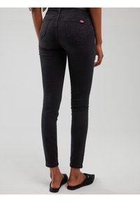 Vedere din spate a unei persoane care poartă blugi skinny negri și papuci slip-on negri, stând pe un podea albă simplă cu un perete alb în fundal.