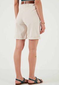 Pantaloni corti beige in cotone con risvolti, dotati di una tasca sul retro. Abbinati a una cintura in pelle marrone e sandali verde teal con cinturini.