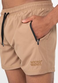 Bañador color beige con cintura elástica, cordón negro y bolsillos laterales con cremallera. Presenta un logo bordado "WRONG FRIENDS" en dorado.