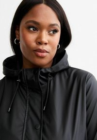 Svart vattentät jacka med huva, med tryckknap stängning, dragsnoddjusteringar och en jämn textur. Enkel, modern design.