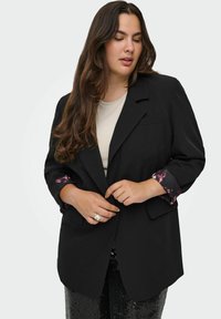 Melns žakets ar pielāgotu dizainu, kas piedāvā zīda ziedu apdruku uz piedurknēm, saskaņots ar neitrālu augšu un izšūtām biksēm.