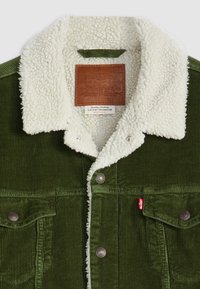 Green corduroy jacket with a white sherpa collar, metal buttons, front pockets, and a leather label at the neck. Textured fabric with visible ribbing.