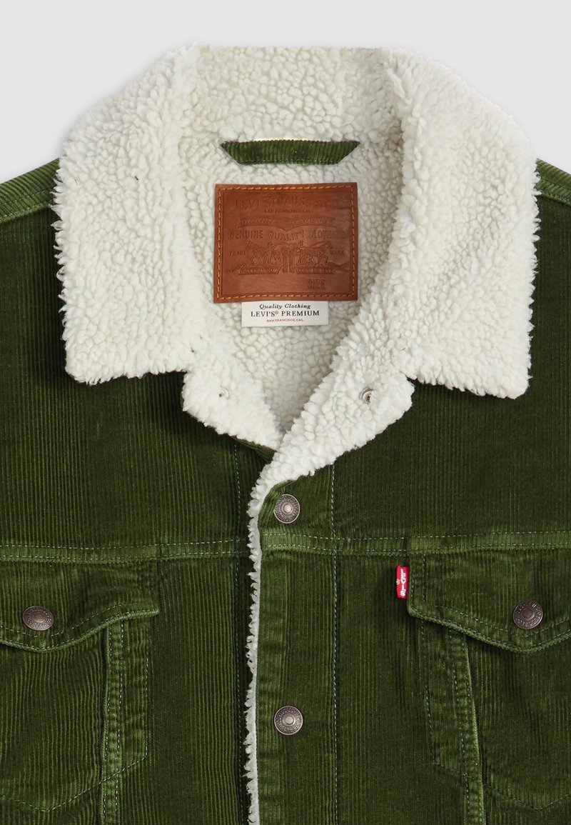 Grüne Cordjacke mit einem weißen Sherpa-Kragen, Metallknöpfen, Fronttaschen und einem Lederetikett am Kragen. Strukturiertes Gewebe mit sichtbarem Rippenmuster.