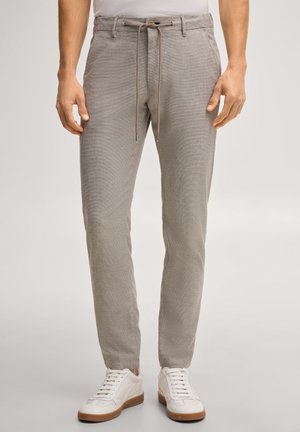 Mężczyzna w jasnoszarych spodniach ze ściągaczem i białych trampkach, stoi z rękami swobodnie opuszczonymi wzdłuż ciała na jednolitym tle.