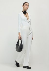 Weißer Langarm-Bodysuit mit tiefem V-Ausschnitt und schwarzem Rosenakzent; kombiniert mit hoch taillierten weißen Hosen und einer schwarzen Handtasche. Schwarze Loafer.