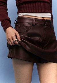 Bordeauxfarbene Ledershorts mit glatter Textur, ausgestattet mit frontalen Taschen und einer mittelhohen Taille, kombiniert mit einem gerippten bordeauxfarbenen Pullover.