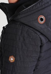 Chaqueta acolchada negra con capucha, con un acento de cuero cerca del cuello y un parche de logo circular en la manga. Material texturizado.