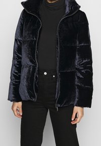 Giacca puffa blu marina in un tessuto lucido e elegante. Presenta un colletto alto e chiusura con zip, con una vestibilità leggermente ampia e tasche laterali.
