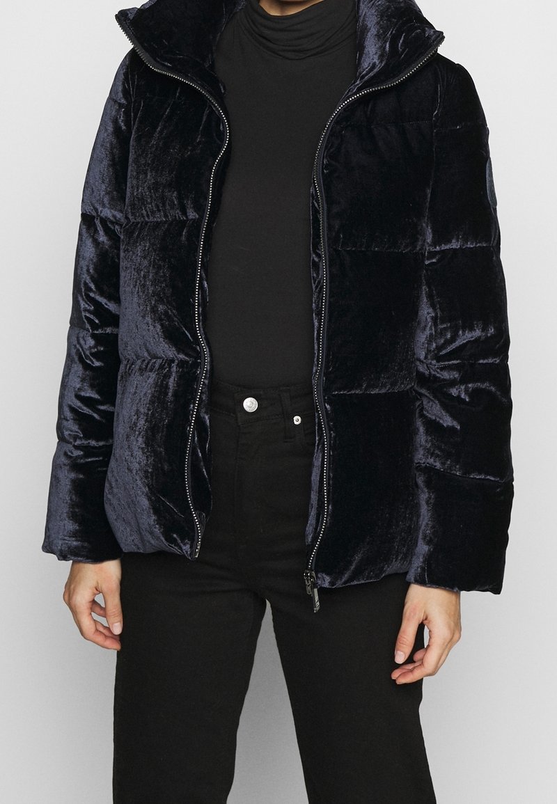 Giacca puffa blu marina in un tessuto lucido e elegante. Presenta un colletto alto e chiusura con zip, con una vestibilità leggermente ampia e tasche laterali.
