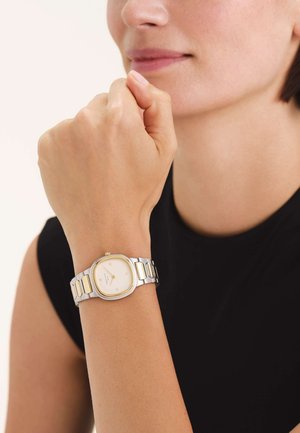 Montre en métal argenté et doré avec un cadran octogonal, un cadran blanc et des maillons rectangulaires sur le bracelet. Portée sur un poignet à la peau claire.