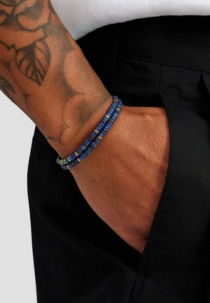 Blåt perlearmbånd med teksturerede runde perler og gyldne accenter, båret på et håndled, delvist synligt i en sort lomme.