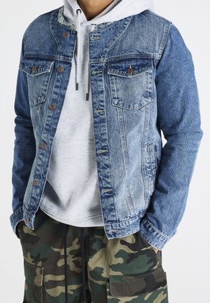 Osoba ubrana w jasnoszary kaptur pod dwukolorową niebieską kurtką dżinsową oraz zielone bojówki w kamuflażu, z rękami w kieszeniach.