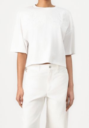 Kobieta ubrana w białą, luźną bluzkę z krótkim rękawem, z subtelnymi haftowanymi kwiatowymi detalami oraz białe spodnie z wysokim stanem.