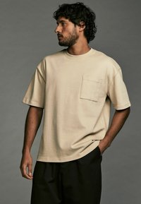 Beige bomulds t-shirt med rund halsudskæring, korte ærmer og en brystlomme, kombineret med mørke bukser. Enkel design og afslappet pasform.