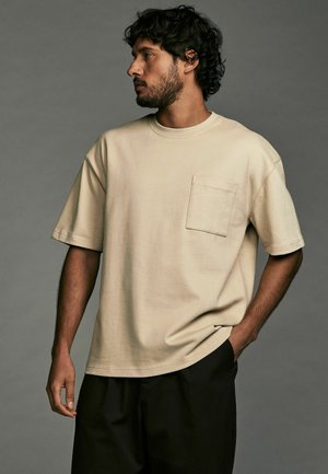 Tricou din bumbac bej, cu guler rotund, mâneci scurte și un buzunar pe piept, asociat cu pantaloni închisi la culoare. Design simplu și croială relaxată.