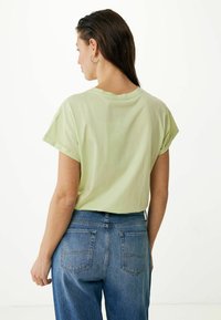Jasnozielona koszula z krótkim rękawem o luźnym kroju; noszona z dżinsami w średnim odcieniu z dwoma tylnymi kieszeniami.