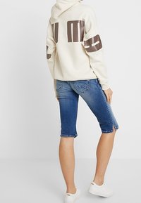 Person som bär en vit huvtröja med bruna grafiker och knälånga blå denimshorts, står med ryggen vänd, vita sneakers på fötterna.