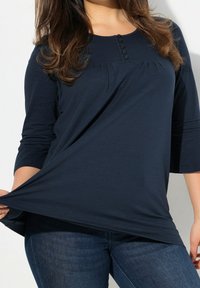 Navy-blaues, langärmliges Oberteil mit Rundhalsausschnitt, Knopfdetails und Raffungen. Kombiniert mit dunklen Jeans. Weicher, fließender Stoff.
