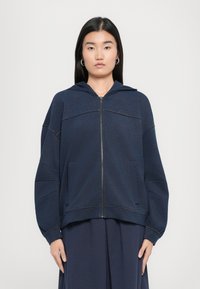 Felpa con zip color navy realizzata in tessuto morbido, caratterizzata da una vestibilità ampia, tasche a marsupio e un cappuccio con cordino e cuciture tono su tono.