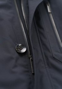 Marinefarbene Oberbekleidung mit einem glänzend schwarzen Knopf und Metallreißverschlüssen auf einem strukturierten Stoff. Nahaufnahme der Details und der Nähte des Kleidungsstücks.