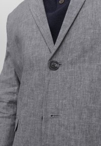 Blazer grigio in lino con un singolo bottone nero, rever a vista e una leggera texture. Indossato sopra un maglione scuro a maglia.