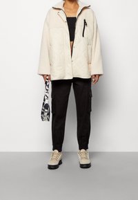 Quiltad ljusbeige jacka med fluffiga ärmslut och en dragkedjeficka, lagd över en svart crop top, tillsammans med svarta cargo-byxor och beige sneakers.