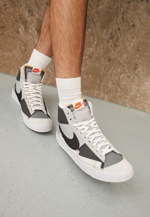 Ben iført hvide sokker og grå, sorte og hvide Nike højtop-sneakers med orange logo, stående på en plettet gulv og brun væg baggrund.