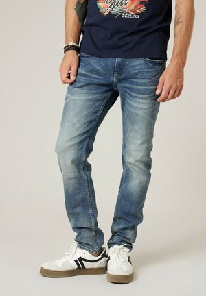 Homme portant un jean bleu délavé, des baskets blanches avec des rayures noires et un t-shirt bleu marine avec un graphisme coloré et un texte.