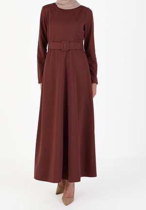 Femme portant une robe maxi à manches longues marron ceinturée, associée à des escarpins beiges à bouts pointus, se tenant devant un fond blanc uni.