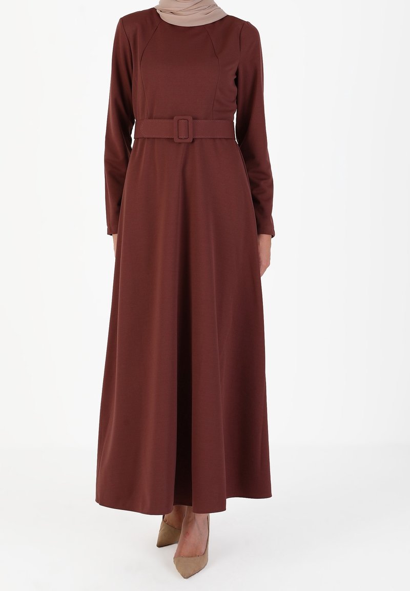 Femme portant une robe maxi à manches longues marron ceinturée, associée à des escarpins beiges à bouts pointus, se tenant devant un fond blanc uni.
