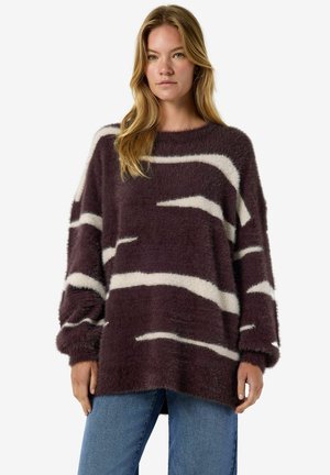 Mulher vestindo um suéter solto e fofo de cor roxo escuro com riscas horizontais brancas irregulares e jeans azul, em pé contra um fundo simples.