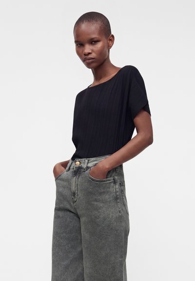 Femme aux cheveux courts portant un haut noir à manches courtes et un jean gris taille haute, debout les mains dans les poches devant un fond uni.