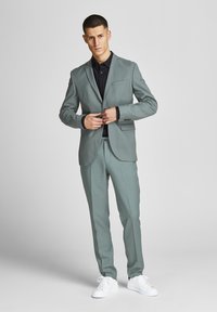 Abito grigio chiaro con giacca strutturata e pantaloni slim-fit. Indossato sopra una camicia nera, abbinato a sneakers bianche. Tessuto morbido, design moderno.