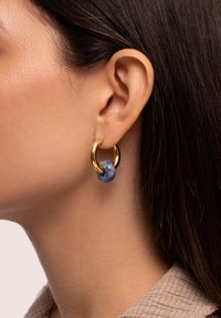 Boucle d'oreille créole en or ornée d'une perle en pierre bleue, portée à l'oreille gauche d'une personne aux cheveux bruns foncés et lisses.