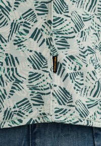 Tela de color claro con un patrón de hojas en verde oscuro, que presenta una etiqueta de marca visible y una superficie texturizada con detalles de costura.