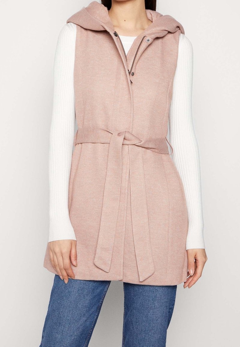 Femme portant une veste sans manches rose clair ceinturée à capuche sur un top blanc à manches longues côtelé et un jean bleu, debout devant un fond clair uni.