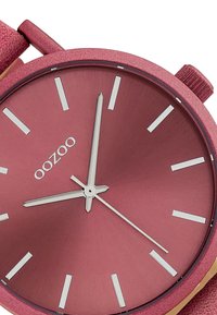 Round watch featuring a rose-colored metal face, white hour markers, and silver hands. Textured pink strap complements the design.