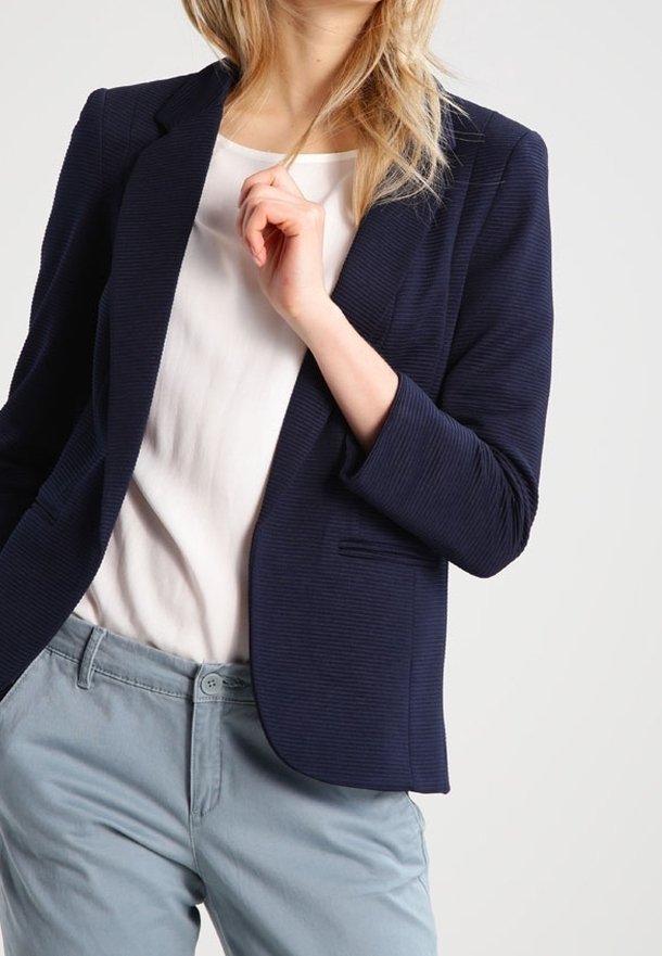 Námořnický žebrovaný blazer s klopami a jedním knoflíkem, nošený přes bílou košili a světle modré kalhoty. Viditelné detaily texturované látky.