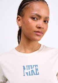 T-shirt couleur crème avec le logo "NIKE" brodé en bleu. Tissu doux avec un col rond classique. Style minimaliste et sans motifs.