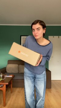 Brązowe pudełko Zalando trzymane przez osobę, z logo. Osoba ma na sobie niebieską koszulę w paski z długim rękawem oraz jasnoniebieskie dżinsy.